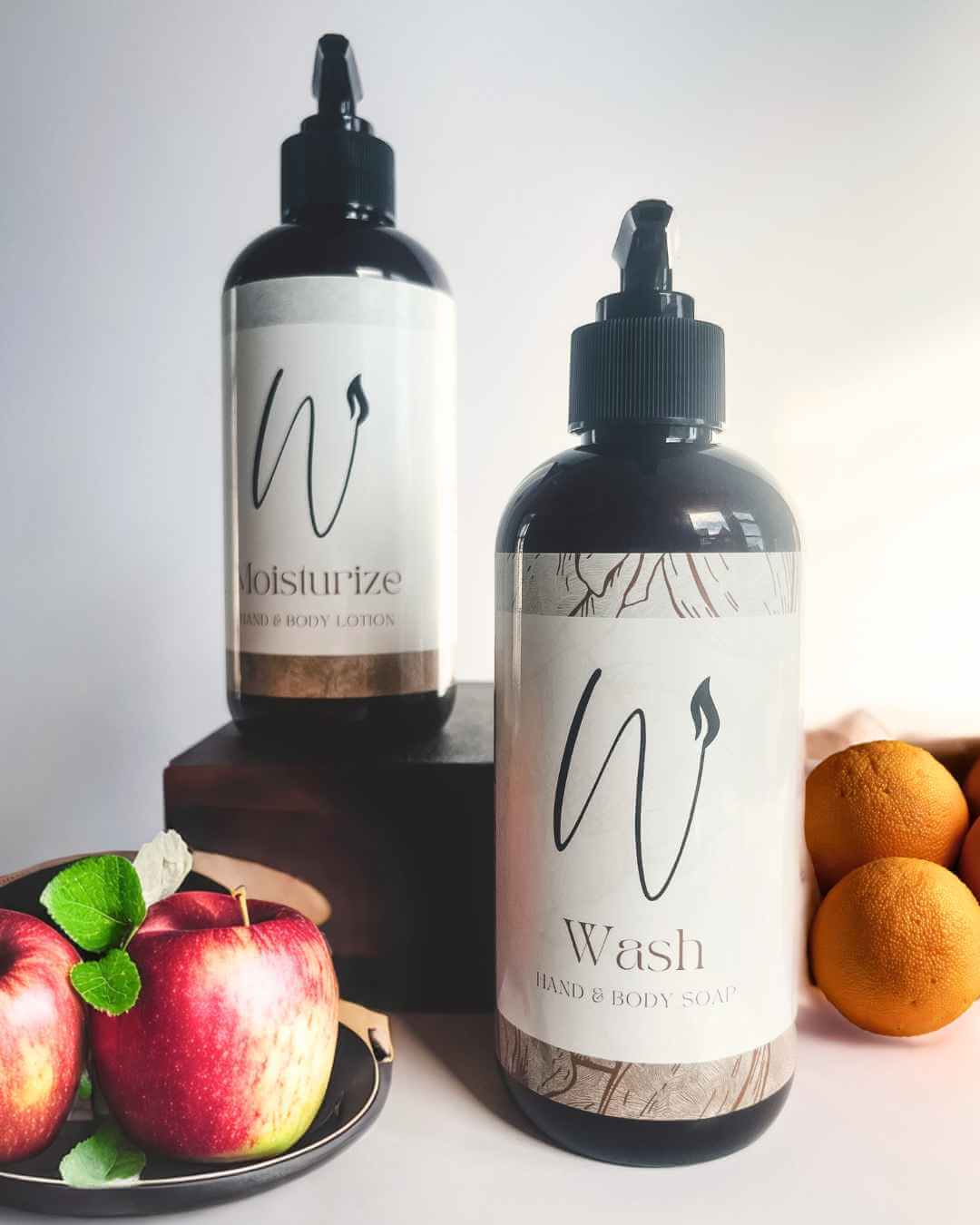 Two bottles of 'W' brand body products with fruits on a white background