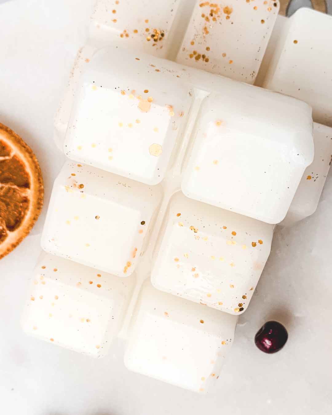 White marshmallows with gold specks on a white surface with a dried orange and cherry.