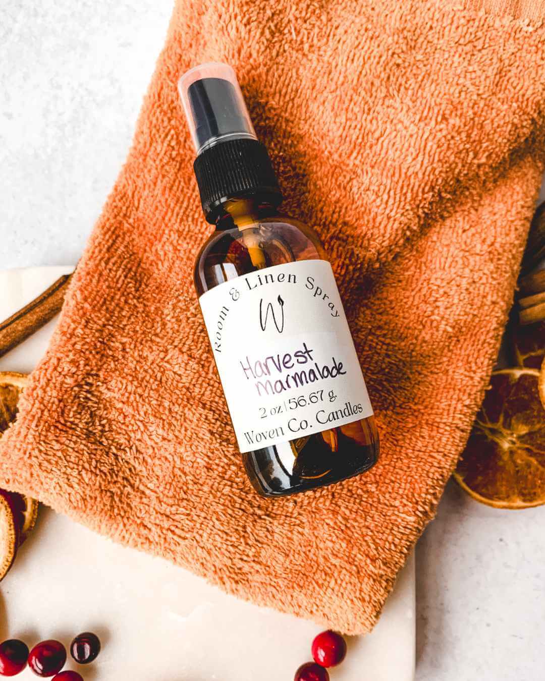Bottle of Harvest Marmalade linen spray on an orange towel with cinnamon sticks and berries.