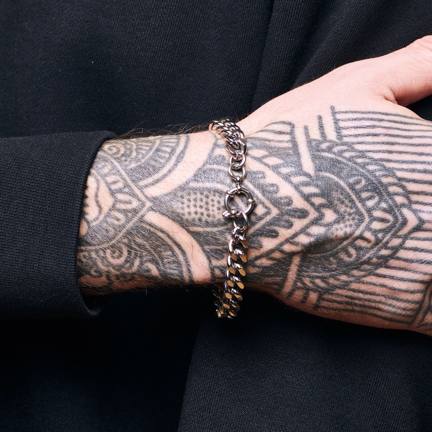 Person wearing a silver chain bracelet on a tattooed arm against a black background