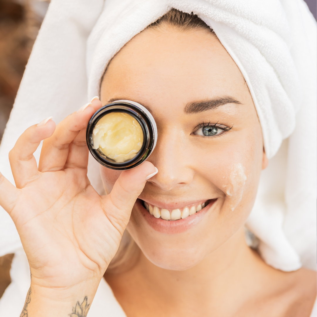 Woman holding a skincare product with a towel on her head, smiling.