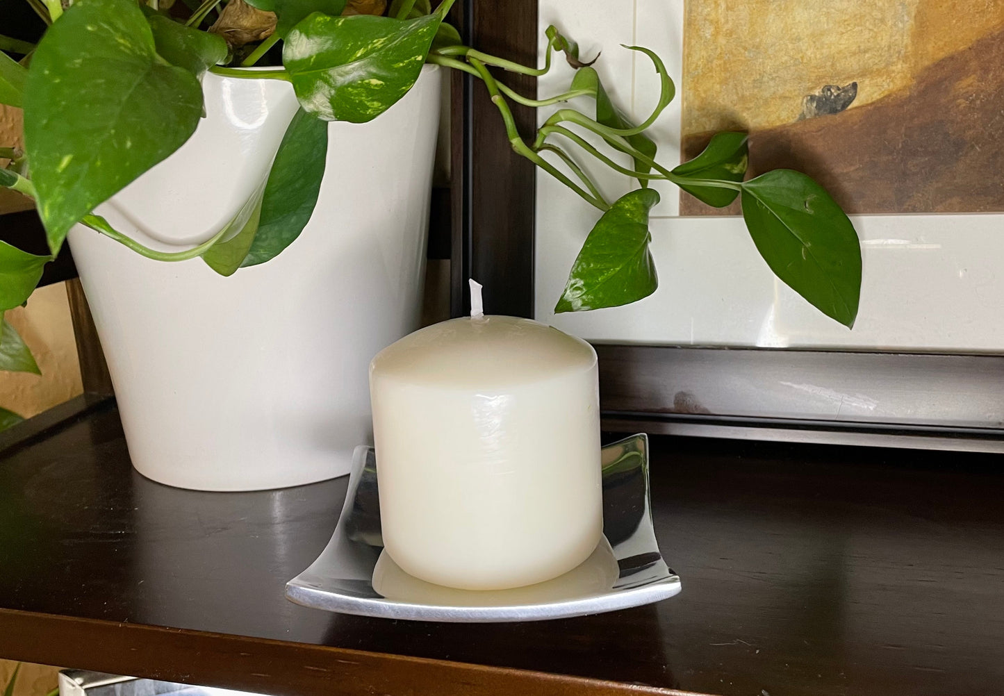 White candle on a reflective surface with a plant and framed picture in the background