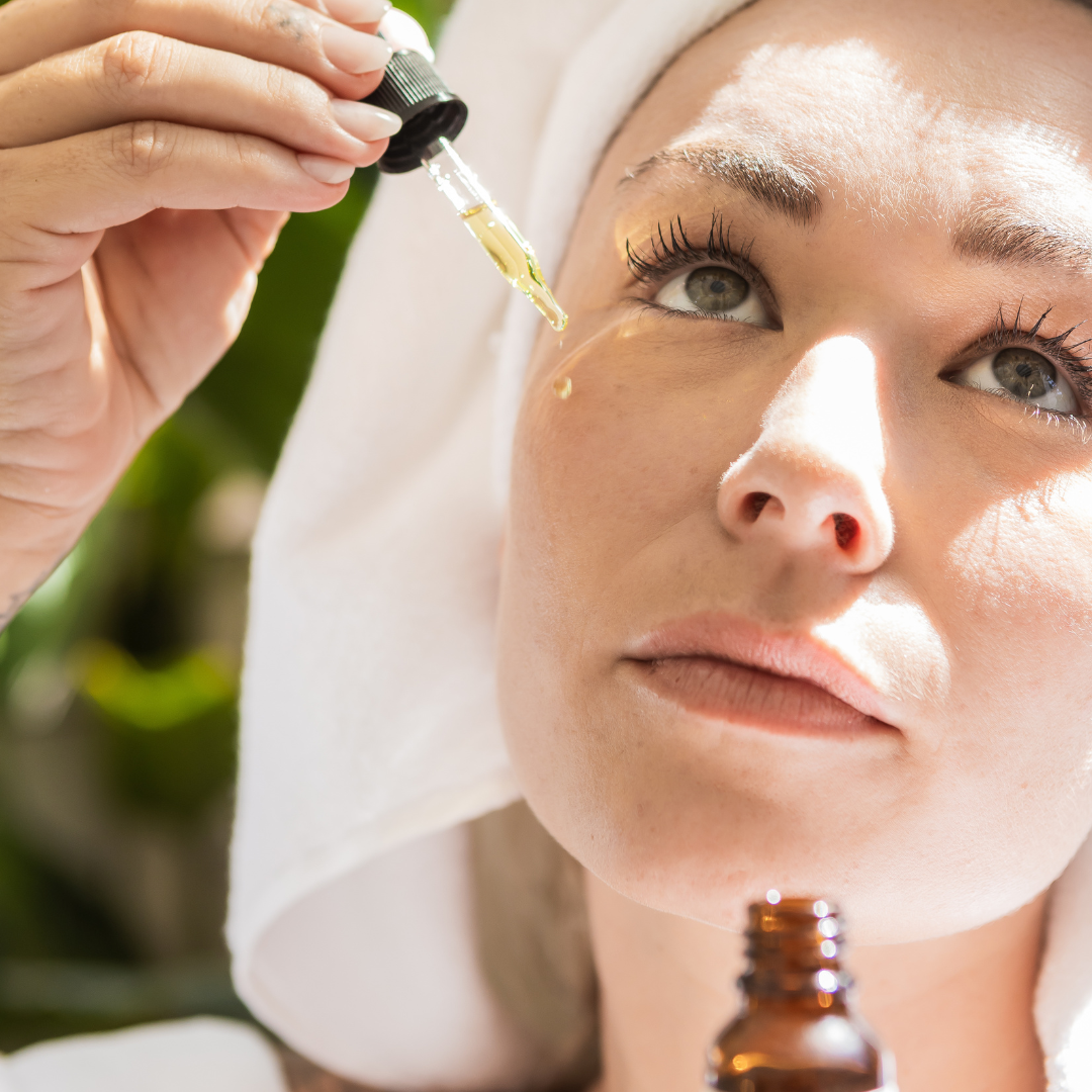 Woman applying a dropper of oil to her face with a towel on her head.