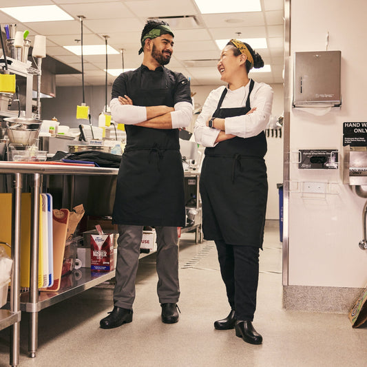 Two chefs in a kitchen, one male and one female, showng off their calzuro shoe for comfort 
