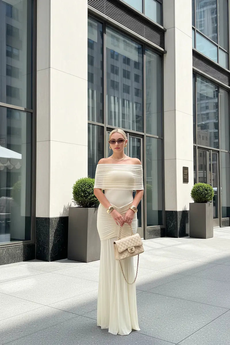 Woman in a white off-shoulder dress standing in front of a modern building.