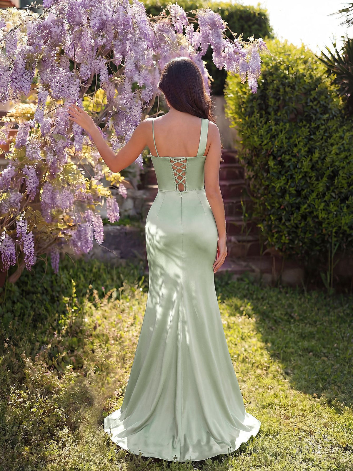 A woman stands outdoors in a garden wearing a mint green satin mermaid-style dress with a lace-up open back. 