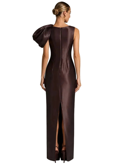 A woman modeling a floor-length dark brown satin gown from the back, featuring a dramatic oversized shoulder detail, a high front slit, and a structured waist.