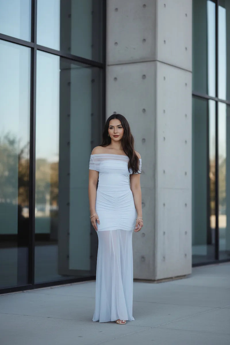 Woman in a white off-shoulder dress standing in front of a modern building with large windows.