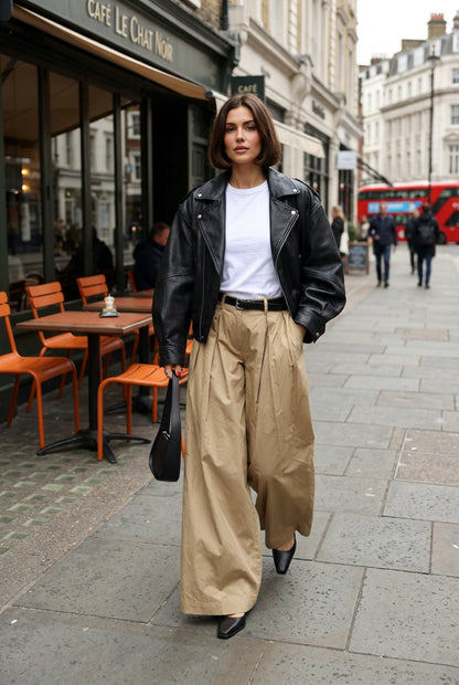 A person wearing wide-leg tan trousers, a black leather jacket, and a white top, holding a black handbag on a city sidewalk.