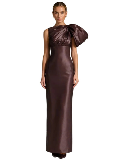A woman modeling a floor-length satin gown in a deep plum-brown color. The dress features a structured, draped shoulder detail and a fitted silhouette, 
