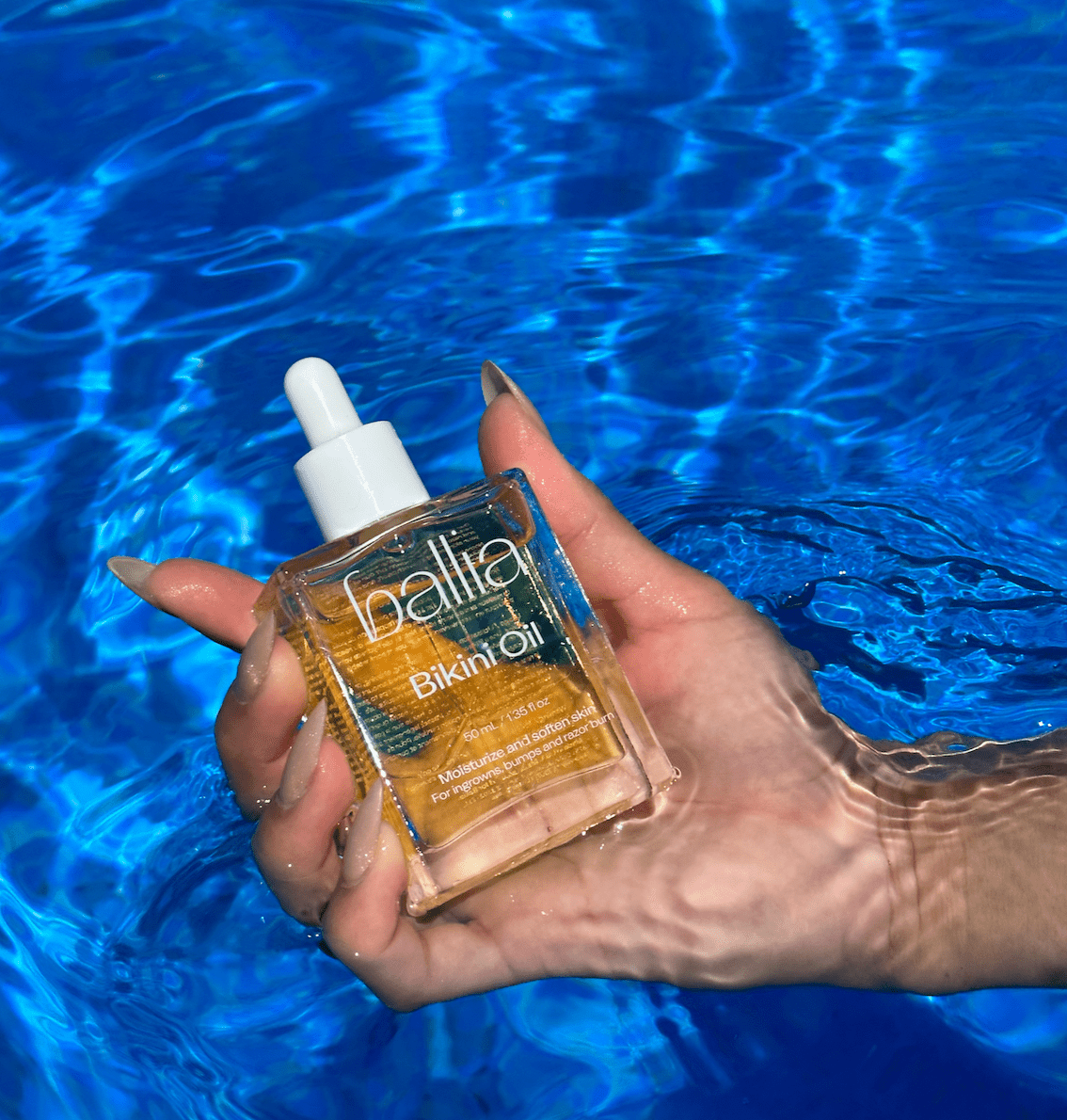 Hand holding a bottle of 'ballia Bikini Oil' with a blue water background