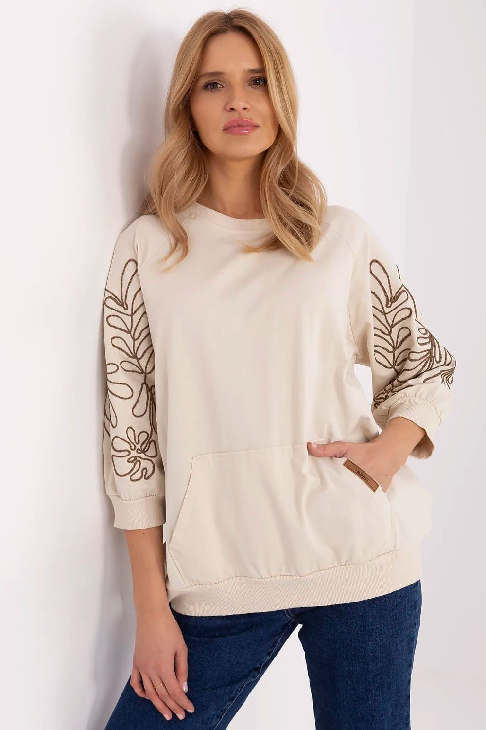 Front view of a model in a beige oversized crewneck sweatshirt with embroidered floral sleeves and blue jeans.