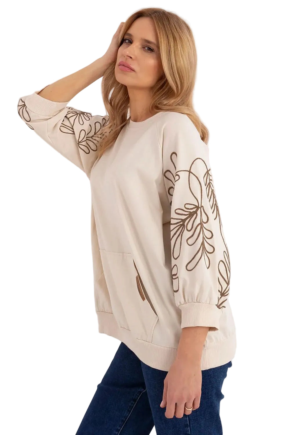 Side view of a model wearing a cream-colored oversized pullover with detailed sleeve embroidery and dark blue jeans.
